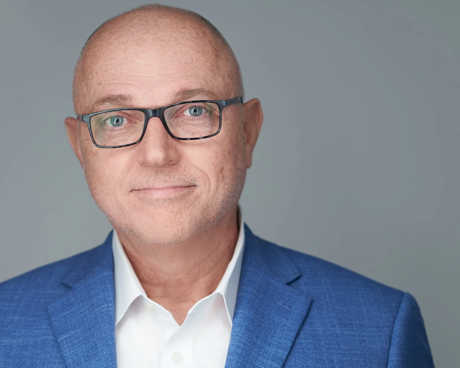 Executive in blue blazer with white open-collar shirt and eyeglasses against gray background — corporate headshot photography