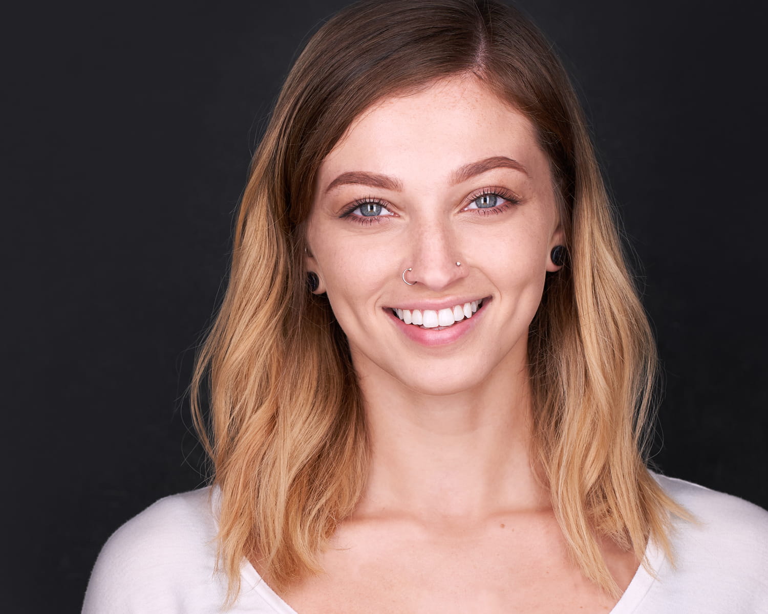 Modeling Headshot Professional modeling headshot of a young woman with blonde hair, natural makeup, and nose piercings, smiling warmly against a dark background in a white top - modern commercial portrait photography for modeling portfolio